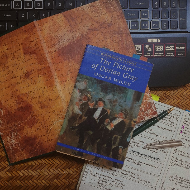 'The Picture of Dorian Gray' by Oscar Wilde on a desk, surrounded by old stationary: notebooks, postcards, and a fountain pen.