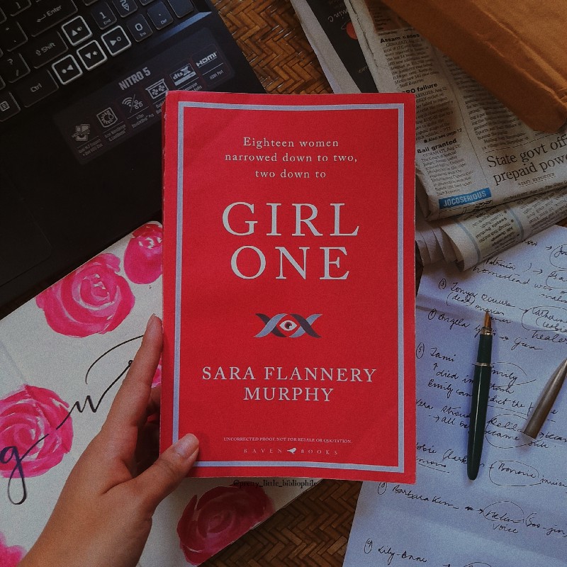 The author holding "Girl One" by Sara Flannery Murphy above a desk with newspapers and a computer underneath.