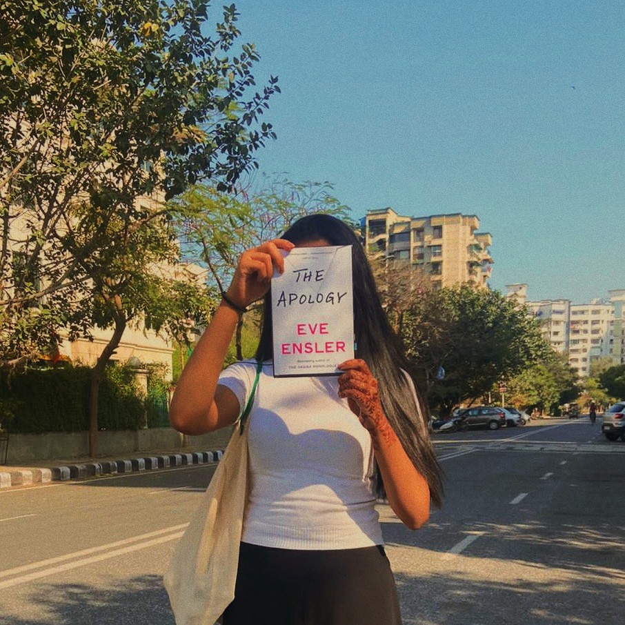 The author holding up a paperback of 'The Apology' by Eve Ensler on a sunny day within a city landscape.