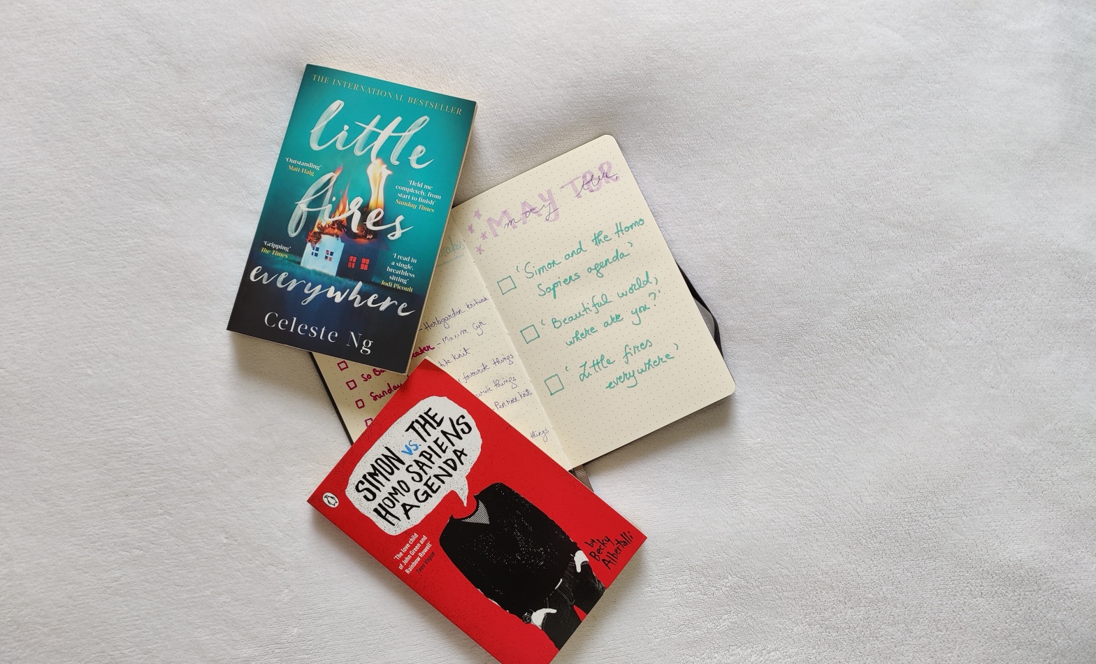 Paperbacks of 'Simon vs the Homo Sapiens Agenda' and 'Little Fires Everywhere' on a white blanket on top of a bullet journal with a May TBR list written on it.