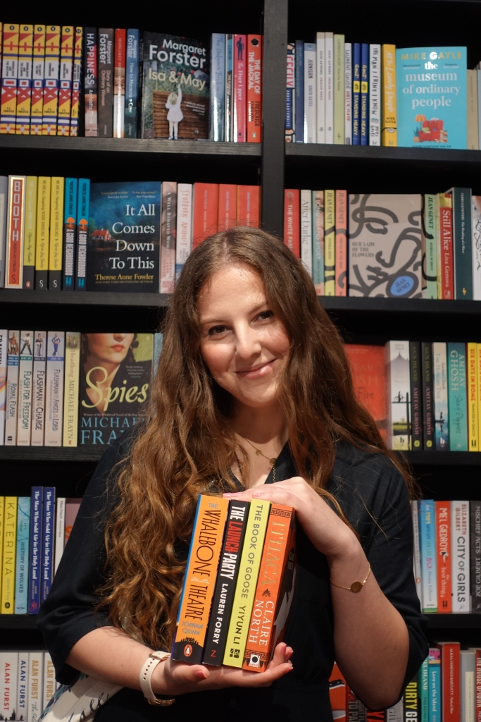 Eliza, a 25 year-old white woman with long chestnut hair, is holding four books and smiling. Behind her, there are bookshelves.